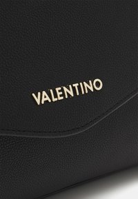 Black textured leather bag with curved flap featuring gold-tone Valentino logo lettering on front.