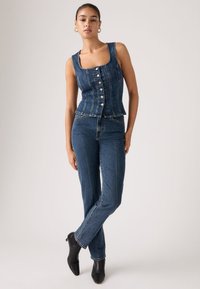 Γυναίκα που φοράει αμάνικο μπλε τζιν πουκάμισο με κουμπιά, ψηλόμεσα τζιν παντελόνια, μαύρα μποτάκια αστραγάλου και χρυσούς κρίκους στα αυτιά, στέκεται με σταυρωμένα πόδια.