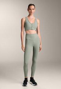 Világoszöld, bordázott rövid top elülső gombos részlettel, amelyhez magas derekú, hozzá illő leggings tartozik. A modellen fekete sportcipő van.