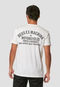 Camiseta de algodón blanca con texto negro impreso en pantalla. El texto dice "DEUS EX MACHINA MOTOCICLETAS PARTES Y SERVICIO" con un acento de estrella.
