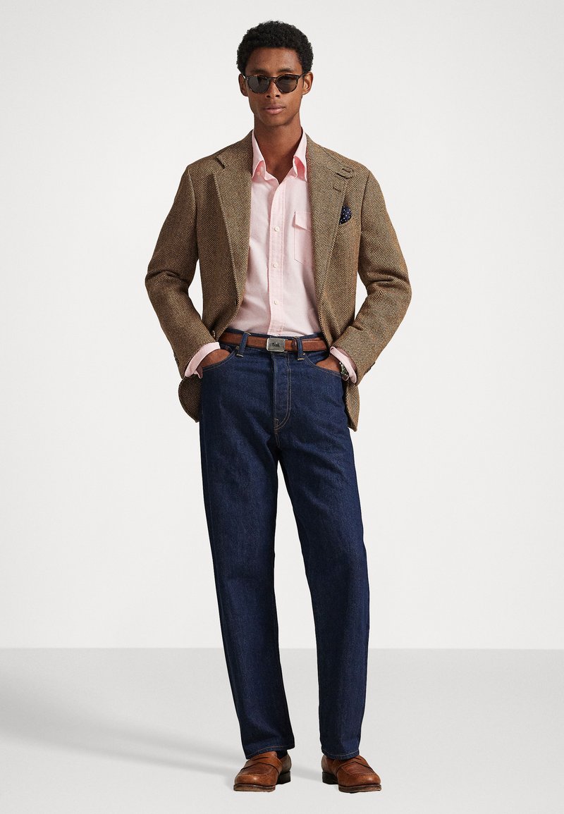 Brown houndstooth blazer, light pink shirt, blue denim trousers, brown leather belt, brown shoes, and sunglasses. Standing pose against a plain background.