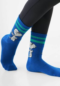 Blaue Socken mit grünen Streifen an der Oberseite zeigen ein Snoopy-Motiv in Weiß und Schwarz. Hergestellt aus geripptem Strickmaterial.