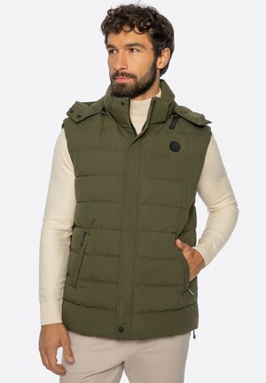 Mężczyzna o kręconych włosach, ubrany w beżowy golf i oliwkową pikowaną kamizelkę bez rękawów, z rękami w kieszeniach.