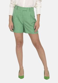 Pantalones cortos verdes texturizados con un diseño plisado, que presentan una cintura alta y bolsillos laterales, combinados con zapatos de tacón alto verdes y una camisa blanca.