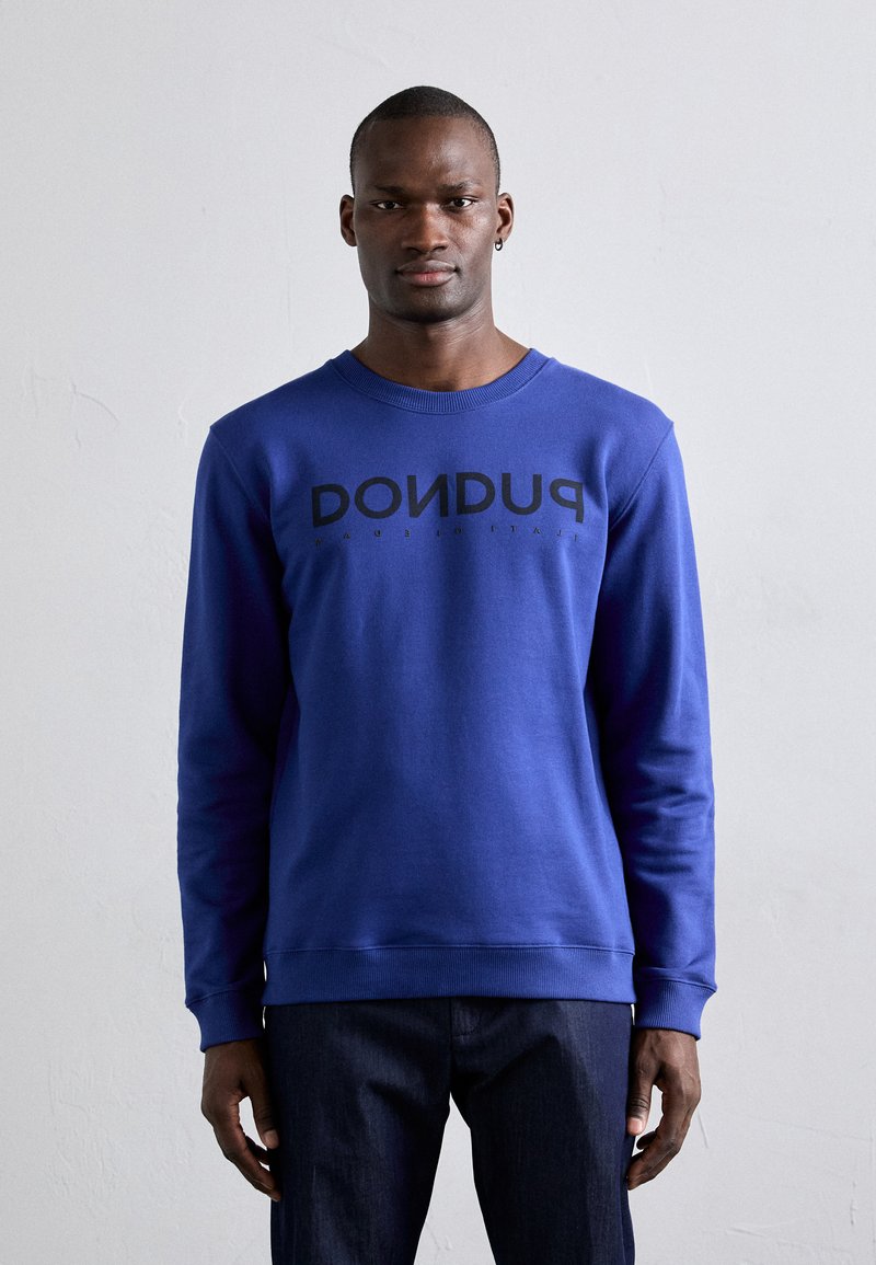 Blue sweatshirt made of cotton with long sleeves, crew neck, and black "PUDU" text prominently displayed on the front. Paired with dark jeans.