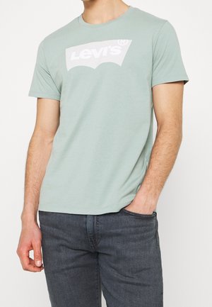 Mand iført en lys grøn Levi's t-shirt og mørkegrå jeans med den ene hånd i lommen mod en enkel baggrund.