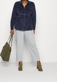 Mörkblå mockaskjorta med knappar, insvängd midja och två bröstfickor, tillsammans med ljusblå vida byxor och olivgröna accessoarer.