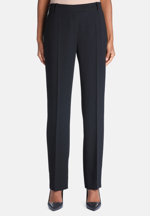 Pantalon droit noir sur mesure porté avec des talons hauts noirs à bout pointu sur une personne debout sur fond blanc.