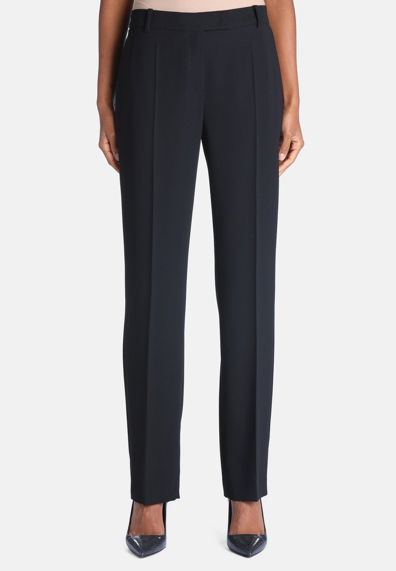 Pantalon droit noir sur mesure porté avec des talons hauts noirs à bout pointu sur une personne debout sur fond blanc.