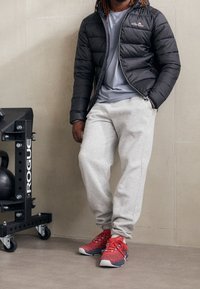 Czarna kurtka puchowa, szary t-shirt, szare dresy, czerwono-niebieskie buty sportowe. Czarny sprzęt gimnastyczny w tle. Gładkie, neutralne tło.