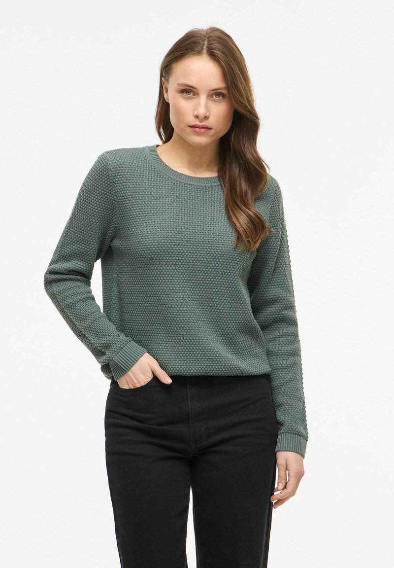 Femme aux longs cheveux bruns porte un pull vert texturé et un pantalon noir, debout, une main dans la poche, sur un fond clair uni.