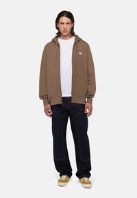 Bruine zip-up hoodie met trekkoorden, voorzien van een klein logo. Gecombineerd met donkerblauwe jeans en gele sneakers. Effen kleur. Casual fit.