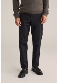 Hombre vistiendo pantalones negros de corte recto, mocasines marrones con borlas y un suéter de punto marrón, de pie contra un fondo beige.