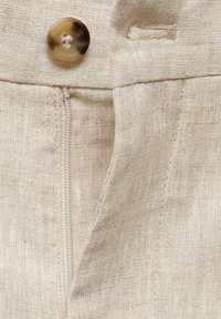 Gros plan sur un tissu beige avec un bouton marron clair au-dessus d'une fermeture éclair partiellement ouverte sur un vêtement.