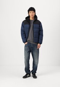 Navy blue puffer jacket with a black yoke, zip closure, attached hood, gray t-shirt, distressed blue jeans, and black sneakers.