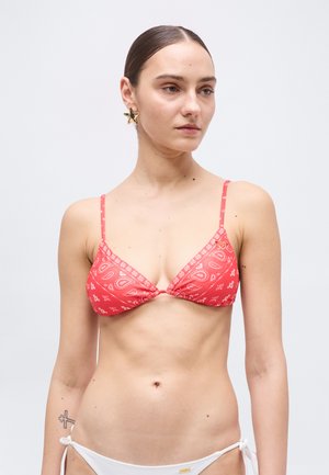 Tânără cu părul dat pe spate poartă un top de bikini roșu cu model paisley în formă de triunghi și slipi de bikini albi cu șnururi laterale, privirea fiind ușor spre dreapta.