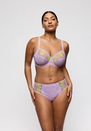 Vrouw met een gemiddelde huidtint die een paarse kanten beha en bijpassende slip met hoge taille en gele bloemrijke borduursels draagt, staand tegen een effen achtergrond.