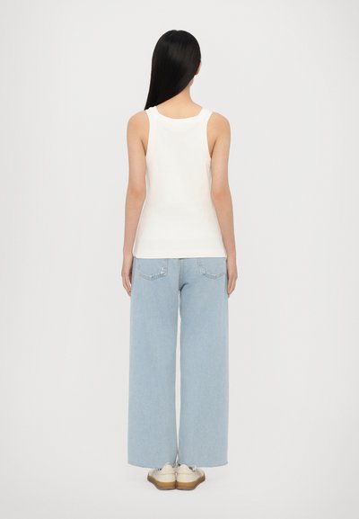 Hvit tanktopp laget av bomull, kombinert med vide lyseblå jeans med baklommer. Personen vises fra bak siden, iført lyse sko.