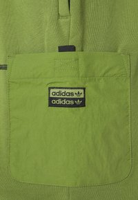 Zelená tkanina s texturovaným povrchem, na kapse s černým vyšitým logem. Design obsahuje posílené detaily šití.