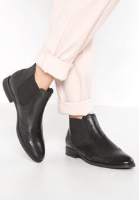 Sorte læder ankelstøvler med elastiske sidepaneler, brogue-detaljer og en lav blokhæl, parret med rullede lyserøde bukser.