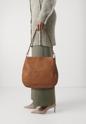 Bruine leren schoudertas met een zachte textuur en gebogen vorm, voorzien van een afneembare schouderband. Gedragen met een lichtgrijze trui en groene broek.