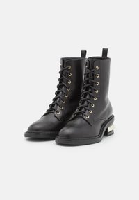 Botas de cuero negro hasta el tobillo con ojales dorados, cordones en la parte frontal, punta redondeada y un detalle de tacón texturizado único. Superficie lisa y diseño robusto.