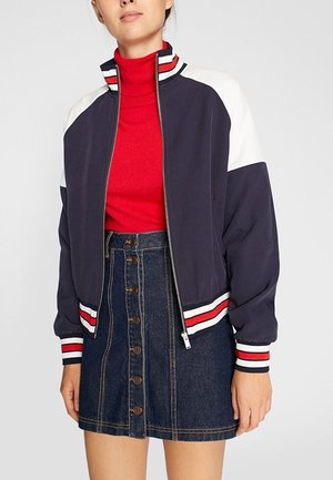 Vrouw die een marineblauw-wit jack met ritssluiting draagt met rood-witte geruite manchetten, een rode coltrui en een donker spijkerrokje met knopen aan de voorkant.