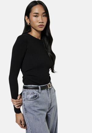 Giovane donna con lunghi capelli neri che indossa un maglione nero a coste, jeans grigi a vita alta e orecchini a cerchio dorati, che posa con una mano dietro la schiena.