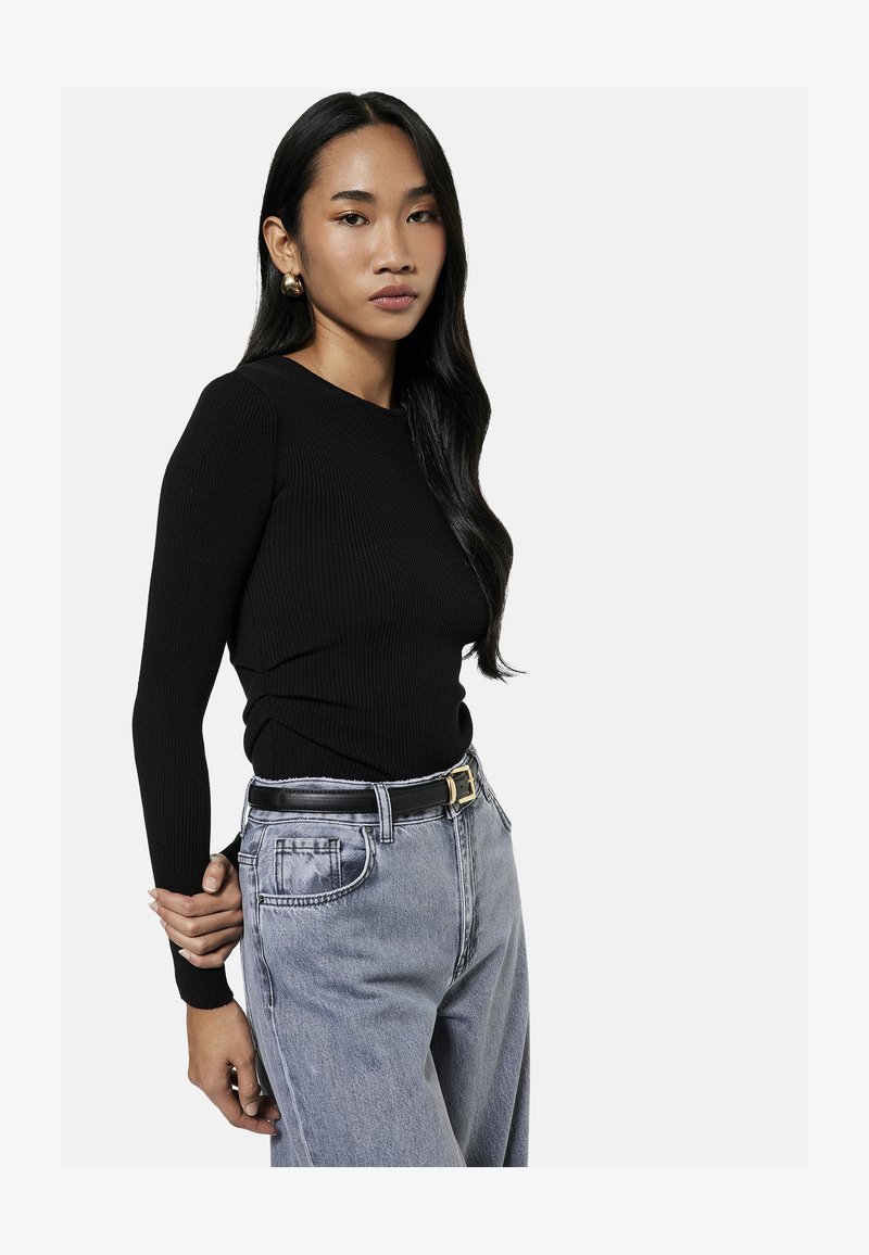 Young woman with long black hair wearing a black ribbed sweater, gray high-waisted jeans, and gold hoop earrings, posing with one hand behind her back.