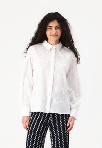 White, embroidered blouse with a button-down collar, long puff sleeves, and floral pattern, paired with navy zigzag patterned pants.