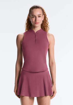 Tânără cu păr creț, îmbrăcată într-un top mov fără mâneci cu fermoar și o fustă asortată, stând în fața unui fundal alb simplu.