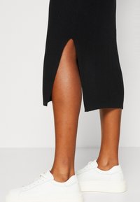 Czarna prążkowana długa sukienka z bocznym rozcięciem, zestawiona z białymi trampkami. Skupienie na fakturze i kontraście kolorów w dolnej części stroju.