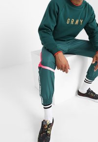Zielona sweatshirt z złotym napisem "GRMY", połączona z turkusowymi dresami z różowymi akcentami po bokach i białymi paskami na mankietach, noszona z ozdobnymi butami.