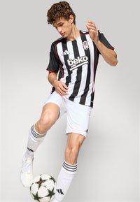 Mustvalge triibuline jalgpallimärk punaste aktsentidega, valgete lühikeste pükstega. Mängija lööb jalgpalli, kannab triibulisi sokke ja kramplikke saapaid.