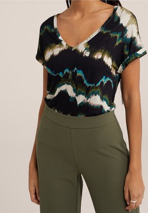 Vrouw die een kortärmelig zwart, turquoise, wit en groen tie-dye V-hals shirt draagt met olijfgroene, hoog getailleerde broek.