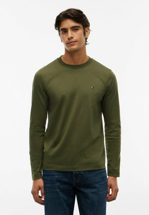 Joven con una camisa de manga larga verde oliva y vaqueros azules de pie contra un fondo blanco liso.