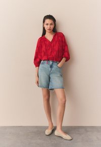 Červená textúrovaná blúzka s výstrihom do V a sťahovacím šnúrkou, skombinovaná s ľadovomodrými rifľovými šortkami s ošúchaným okrajom. Outfit dopĺňajú béžové pletené baleríny.