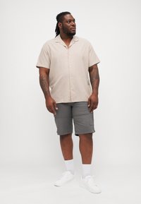 Camicia a maniche corte beige chiaro con bottoni, abbinata a pantaloni cargo grigi. Le sneakers bianche e calzini alla caviglia completano l'outfit.