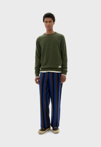 Suéter de punto verde con mangas largas, combinado con pantalones de pierna ancha a rayas verticales en azul y marrón, usados con zapatillas beige.