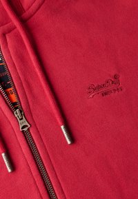 Felpa rossa con zip e cappuccio, con logo "SuperDry" ricamato, hardware della zip in metallo e dettagli con cordini. Texture liscia con un design casual.