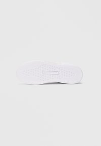 Fehér sneaker talp, textúrált tapadási mintával és a középen elhelyezett Tommy Hilfiger logóval az alján, sima háttér előtt.