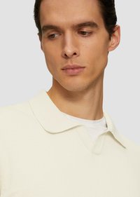 Jasno kremowa, dzianinowa koszulka polo o miękkiej fakturze, z klasycznym kołnierzykiem i dekoltem w kształcie V, noszona na białej koszulce pod spód.