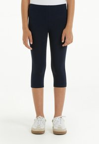 Kapitánové legíny v námořnické modři vyrobené z elastického materiálu, s hladkým povrchem a elastickým pasem. Nosené s bílými sportovními teniskami.