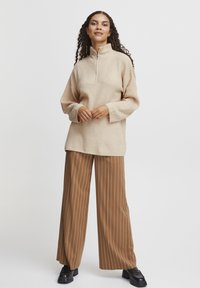 Beżowy sweter na zamek z prążkowaną fakturą, w połączeniu z brązowymi spodniami w paski o szerokich nogawkach i czarnymi butami na platformie. Model stoi na tle jednolitego tła.