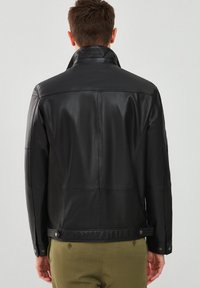 Giubbotto di pelle nera con colletto alto, cuciture visibili e bottoni ai polsi e alla vita. Struttura liscia, design aderente.