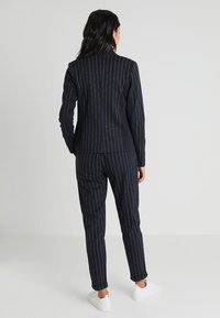 Marinblå randig kostymjacka och byxor i texturerad tyg, med vertikala ränder i ljusa accentfärger och vita sneakers.