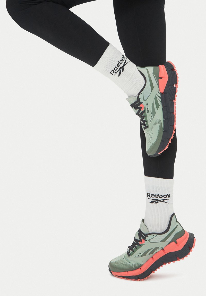 Svetlo zelena športna čevlja s črnimi in koralnimi poudarki; zgornji del iz mrežastega materiala, teksturiran podplat in črne vezalke. Bele nogavice z logotipom Reebok.