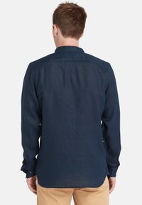Maglietta a maniche lunghe blu navy realizzata in tessuto leggero; presenta un colletto, polsini con bottone e un orlo dritto con una vestibilità leggermente rilassata.