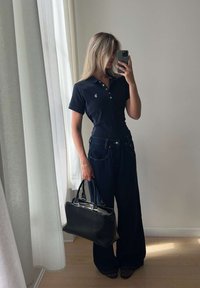 Kvinne iført svart poloshirt og vide jeans, holder en svart håndveske og tar en speilselfie i et lyst rom.