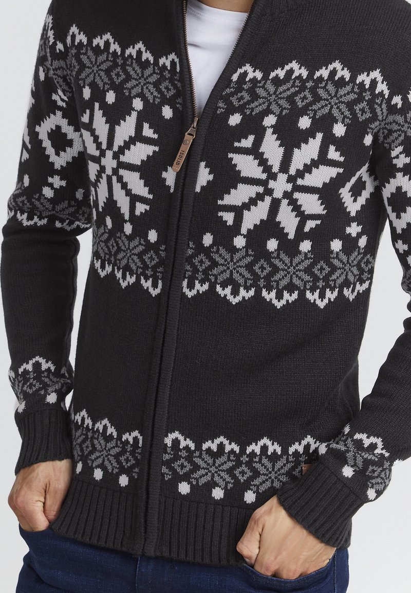 Pull noir en maille à fermeture éclair avec de grands motifs de flocons de neige blancs et gris et motifs géométriques, porté sur une chemise blanche.
